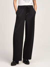 A person wearing black wide-leg drawstring pants with a white graphic T-shirt and black platform shoes, photographed against a plain light gray studio background.
