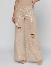 A model wears a pair of wide-leg sequined pants in a champagne beige color, featuring distressed rips at the knees. The pants are covered in shimmering sequins and paired with a beige top and beige pointed-toe heels against a light gray studio background.