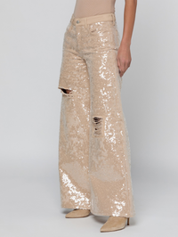 A model is shown from the waist down wearing wide-leg, distressed jeans covered in shimmering champagne beige sequins, paired with beige pointed-toe heels against a light gray studio background.