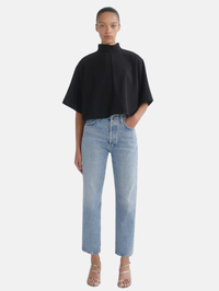 A model wearing a black short-sleeved mock-neck top and light-wash blue straight-leg jeans, standing against a light gray studio background.