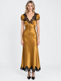 A woman modeling a floor-length satin slip dress in a rich mustard-gold color with black lace trim at the neckline, sleeves, and hem, paired with black heels against a plain white studio background.