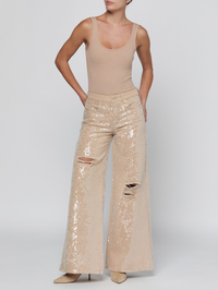 A model wearing beige sequined wide-leg pants with distressed knees and a matching beige ribbed tank top, standing against a light gray studio background.