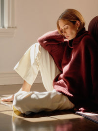 Seated model in the SABLYN Scarlett Slouchy Mock Neck Sweater in Cabernet with ivory pants in soft window light