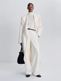 Full-length view of the SIMKHAI Carrie Fringe Coat in Frost worn open over winter white trousers.