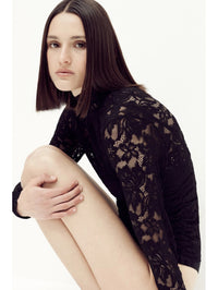 Seated side view of a model in the SIMKHAI Zurie Lace Bodysuit in Black showing the floral lace sleeve.