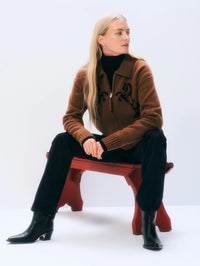 Seated model in the White & Warren Cashmere Blend Horse Bomber in Umber/Black with black denim and boots.